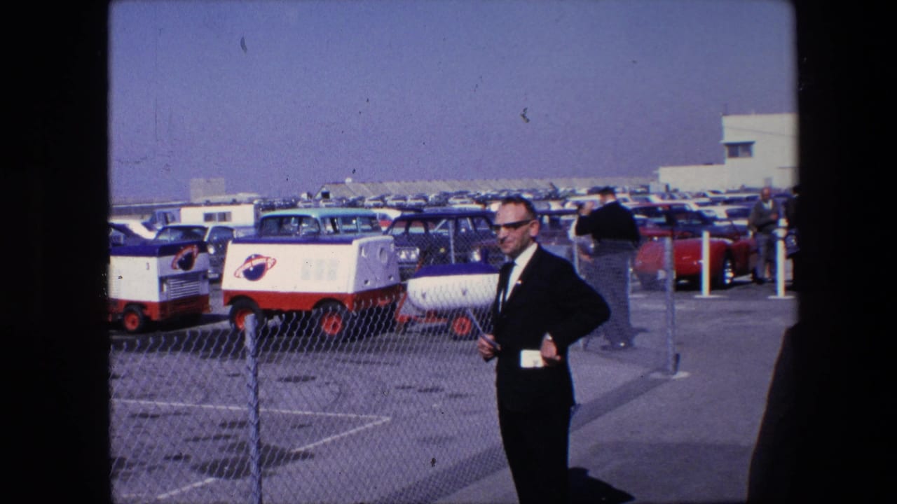 Suited man with sunglasses stands with one hand at his side beside a chain-link fence, with parked cars and red-and-white carts in the background.