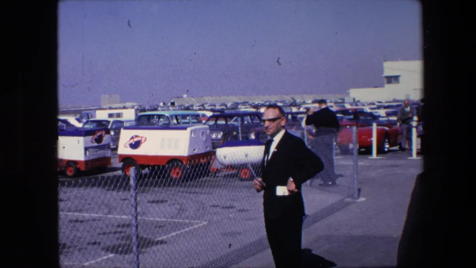 1967 8mm frame of a man in a dark suit and sunglasses standing near a fenced parking area, with rows of older vehicles and red-and-white carts behind him.