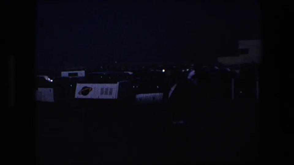 Dim 8mm frame at dusk showing the same parking area in silhouette, with faint outlines of cars, carts, fence, and a low building form.
