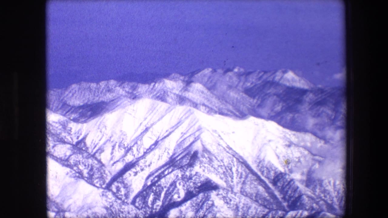 1967 8mm frame showing snowy mountain ridges and distant peaks under a blue sky, with purple-blue film aging and dust visible in the scan.