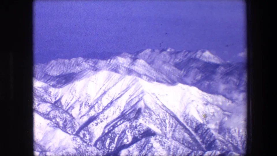 1967 8mm frame showing snowy mountain ridges and distant peaks under a blue sky, with purple-blue film aging and dust visible in the scan.
