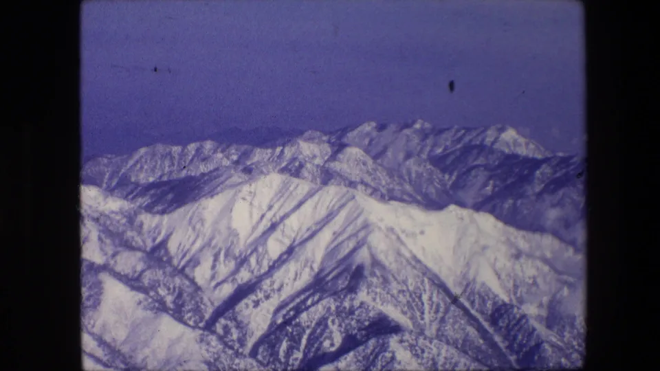 Snow-covered mountain range seen from an elevated or aerial viewpoint, with layered peaks, dark slope lines, and aged 8mm color shift.