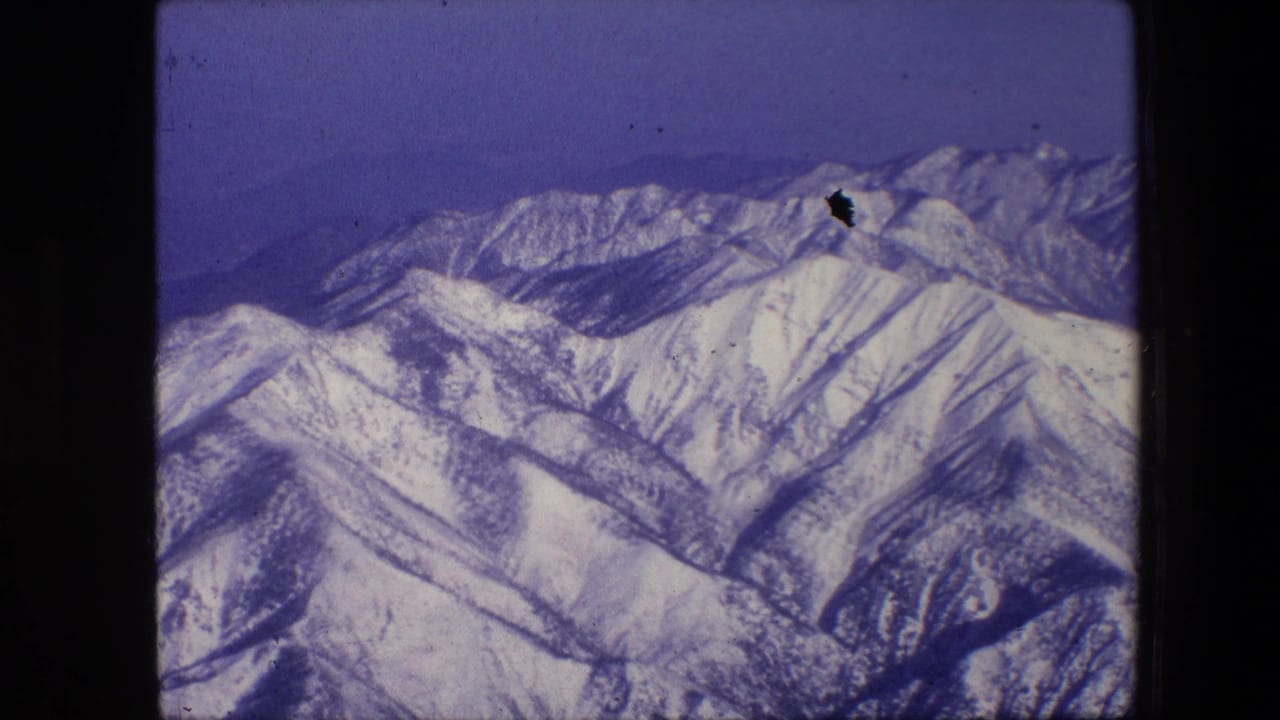 1967 8mm frame showing snow-covered mountain ridges and distant peaks under a blue sky, viewed from an aerial or elevated position.