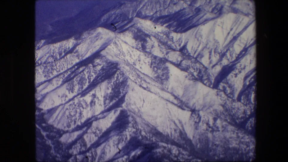 1967 8mm frame of snow-covered peaks and layered ridgelines, with blue-purple film color and visible scanned frame borders.