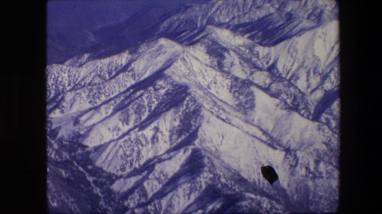 1967 8mm aerial view of snow-covered mountain ridges under a deep blue sky, with visible gate edges and film specks.