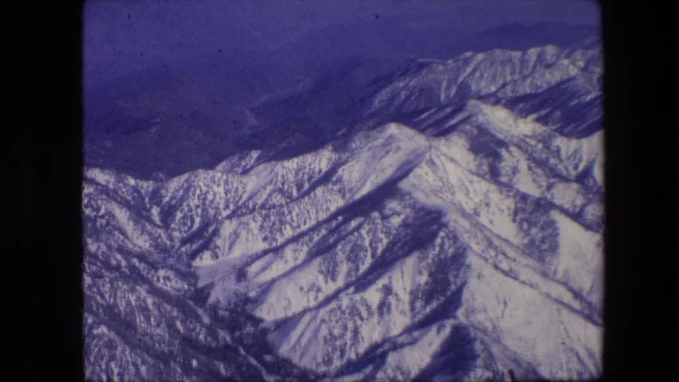 1967 8mm aircraft-view shot of snow-covered peaks and shadowed ridges with a blue color cast and soft home-movie texture.