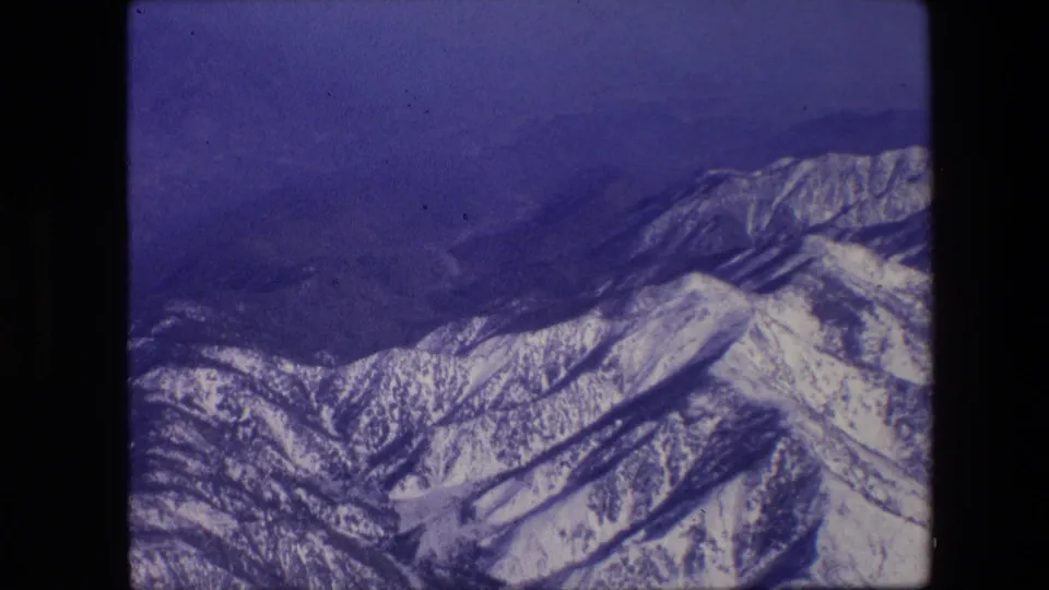 1967 8mm aerial frame looking across snow-streaked mountain slopes and layered ridges, with blue haze and visible film grain.