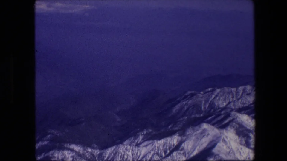A snowy mountain range fills the lower frame beneath a dim blue-purple sky, with dark film and aircraft-window borders visible around the view.