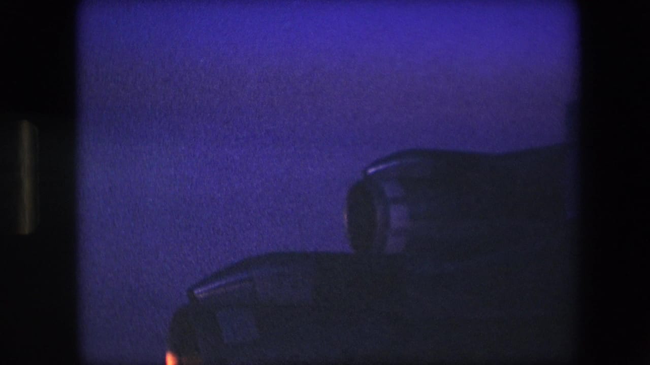 1960s 8mm-style close-up of dark aircraft engine housings with a faint warm glow at the lower left and heavy blue low-light grain.
