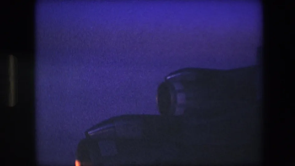 1960s 8mm-style close-up of dark aircraft engine housings with a faint warm glow at the lower left and heavy blue low-light grain.