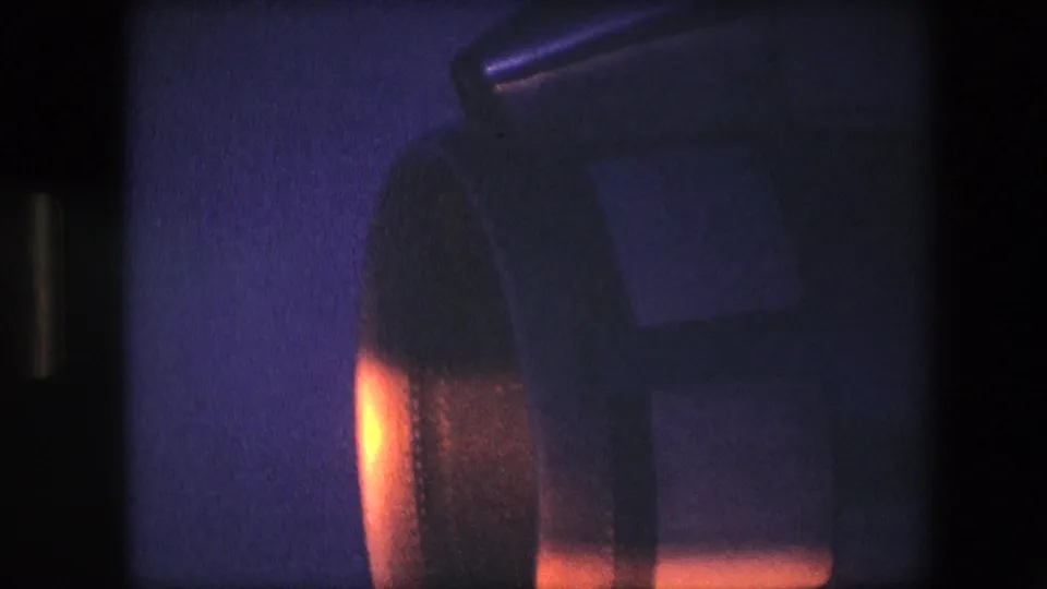 1967 archival 8mm frame showing a close view of a turbine-like aircraft component under orange light against a blue-purple low-light background.