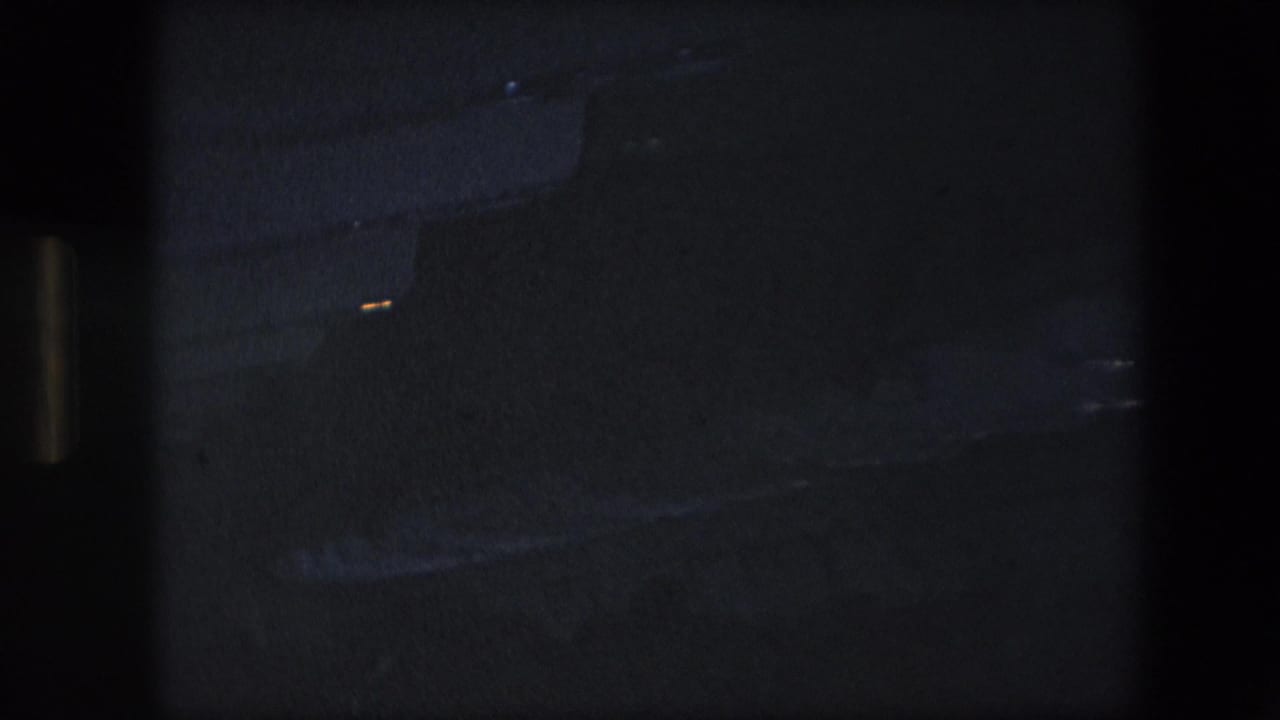 Dark 8mm frame from an airliner interior, showing a mostly underexposed cabin area with faint ceiling and aisle details.