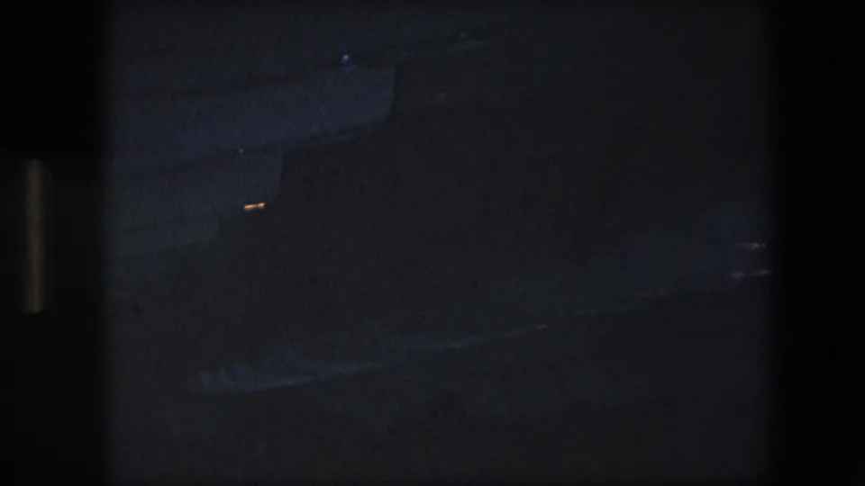 Dark 8mm frame from an airliner interior, showing a mostly underexposed cabin area with faint ceiling and aisle details.