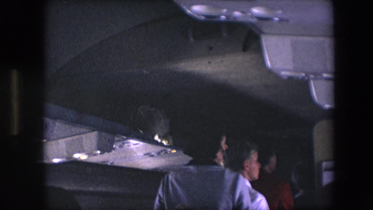 Women standing or moving in a dim airplane aisle beneath overhead compartments.