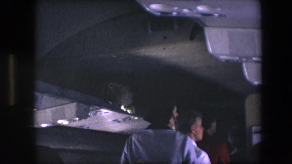 1967 8mm airliner cabin view with women visible in the aisle under overhead compartments and low interior lighting.