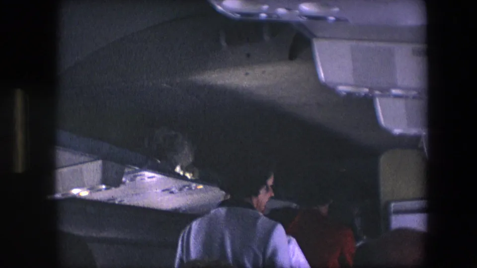 Handheld 8mm view of a woman in a light-colored jacket seen from behind in a narrow airliner aisle, with overhead bins and seats nearby.