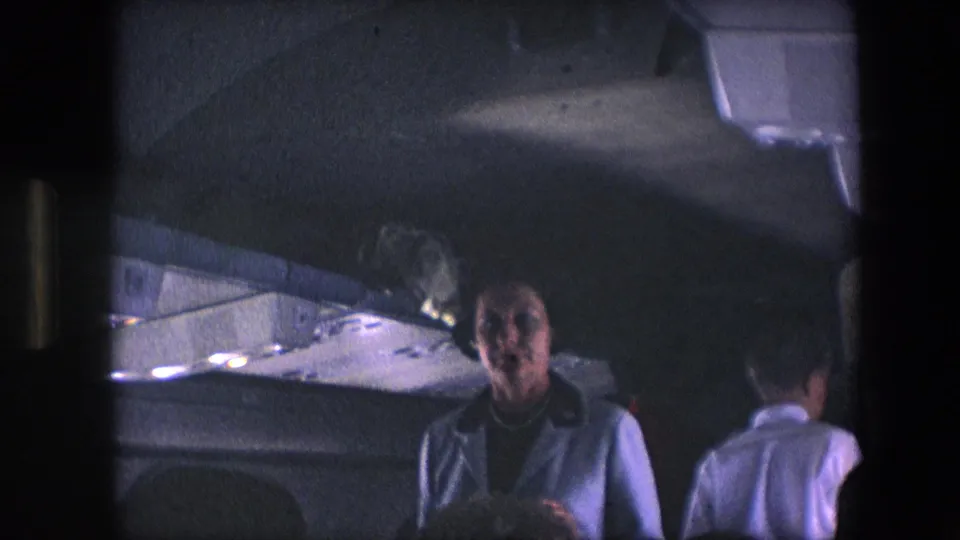 1967 airliner interior frame showing two women in the aisle, including one woman facing toward the camera under dim overhead cabin lighting.