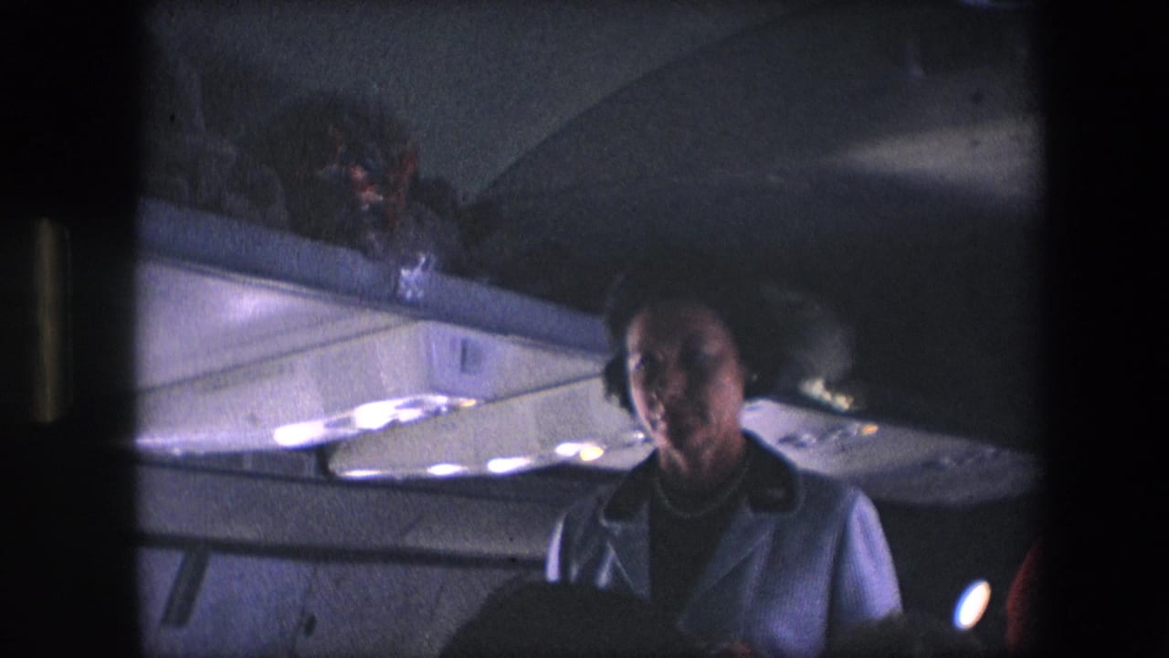 A flight attendant in a light uniform stands in a dimly lit commercial aircraft cabin, with overhead compartments and ceiling lights visible in archival 8mm footage from the 1960s metadata context.