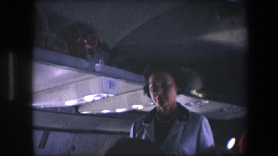 A flight attendant in a light uniform stands in a dimly lit commercial aircraft cabin, with overhead compartments and ceiling lights visible in archival 8mm footage from the 1960s metadata context.