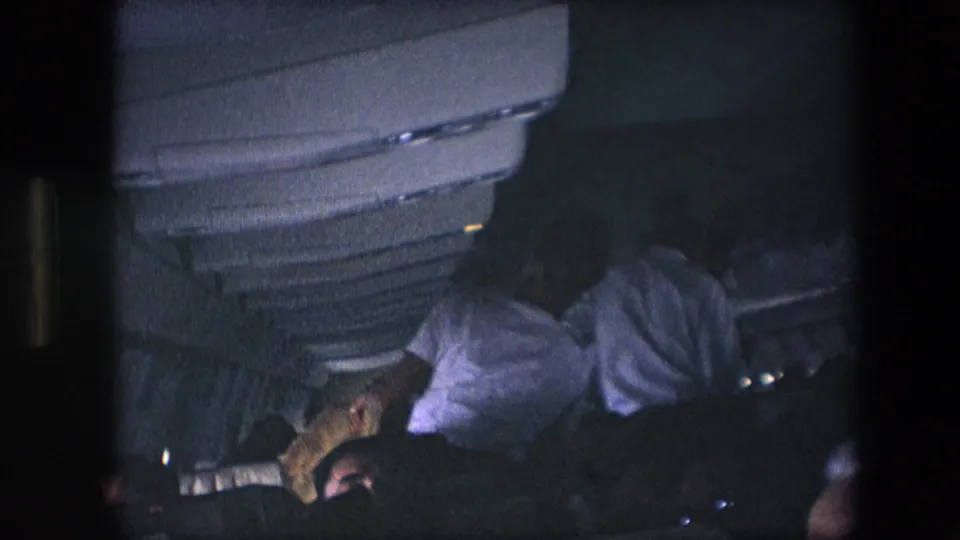 Seated passengers are visible beneath overhead compartments in a dim aircraft cabin, with one person leaning or turning while the handheld 8mm camera records from a nearby seat.