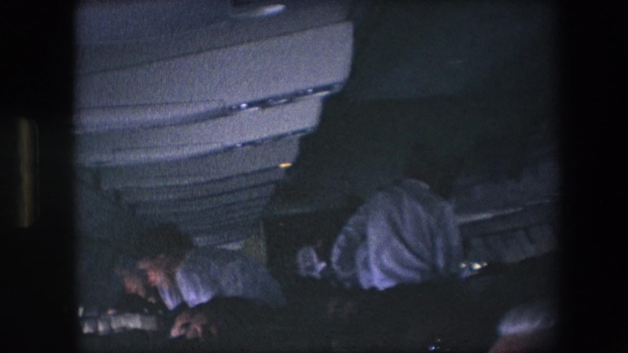 1967 archival 8mm frame of passengers inside a dark commercial airplane cabin, with overhead bins above and travelers settling into seats.
