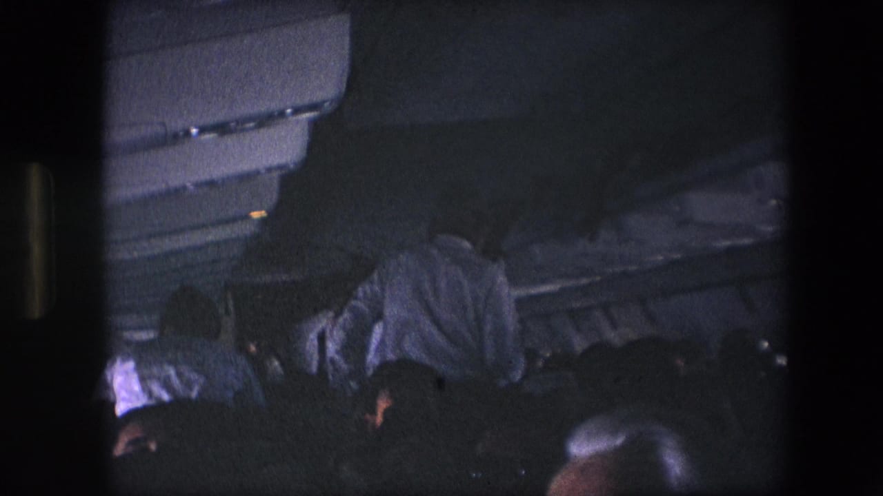 Several passengers sit or lean forward in a dark airplane cabin with overhead compartments visible.