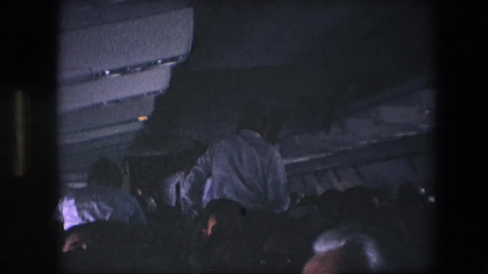Archival home-movie frame showing passengers in a low-lit airplane cabin, with a man in a light shirt leaning near the aisle beneath overhead compartments.