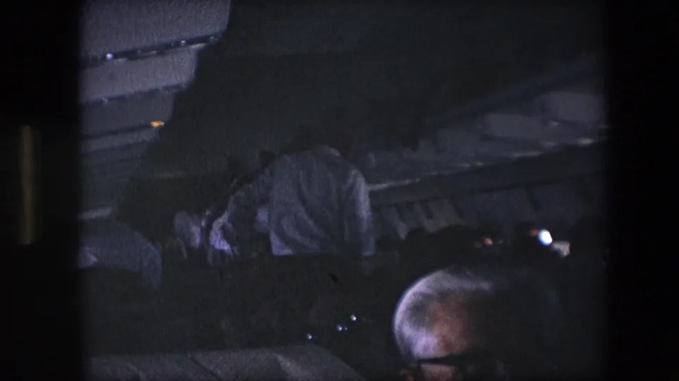 Low-light 8mm frame from inside a commercial airplane cabin, showing a passenger bent forward among rows of dark seats and overhead storage bins.
