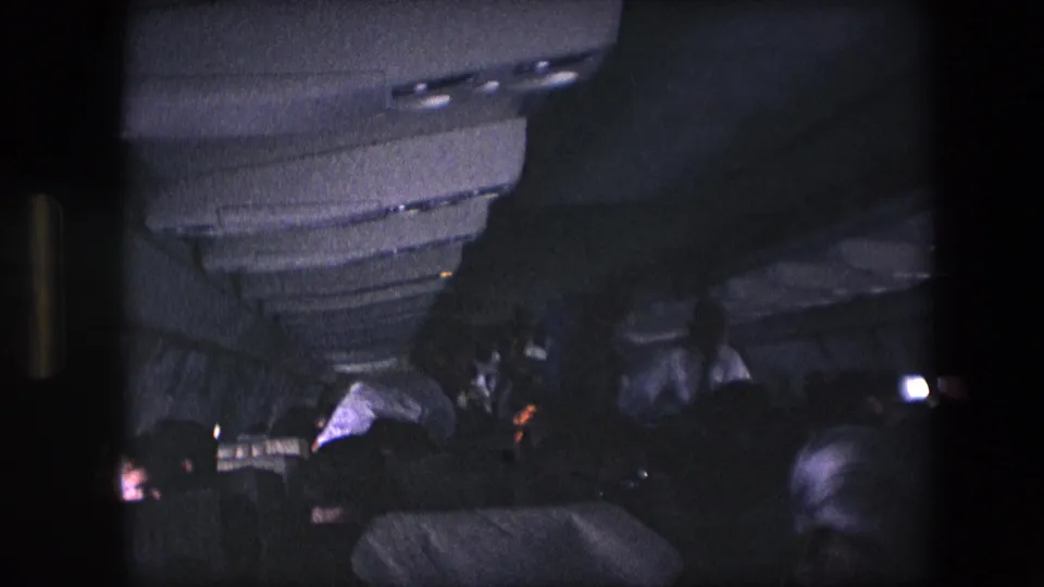 1960s archival airplane-cabin frame with passengers seated or leaning forward under overhead storage compartments in a dark home-movie exposure.