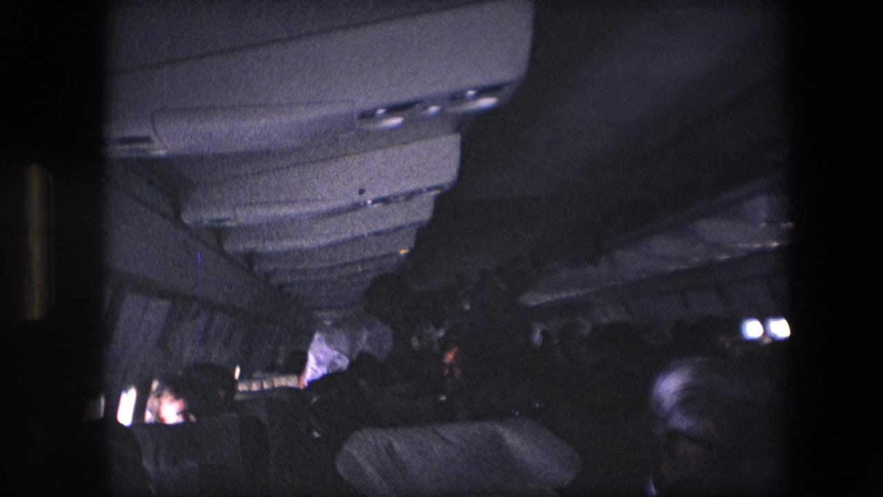 1967 8mm home-movie frame showing a dark airplane cabin interior with overhead panels, seat backs, and passengers under low cabin light.
