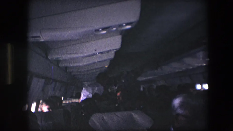 1967 8mm home-movie frame showing a dark airplane cabin interior with overhead panels, seat backs, and passengers under low cabin light.