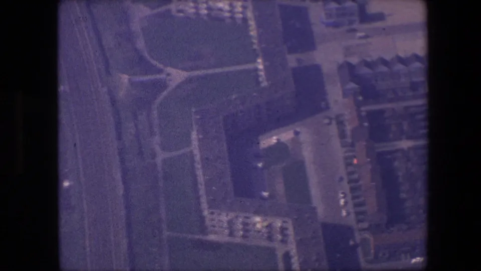 1967 home-movie aerial view from an aircraft window, showing roads, buildings, and a large dark-roofed structure below.