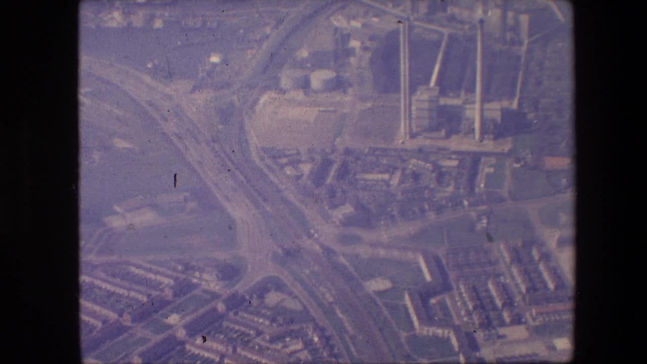 1967 8mm aerial frame, per metadata, showing a New Jersey industrial area with curving highway ramps, a broad waterway, large buildings, and tall smokestacks under a purple color cast.