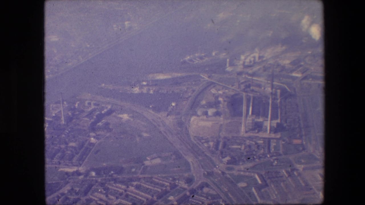 Aerial 8mm frame from the supplied 1967 New Jersey clip showing highway approaches crossing near a wide waterway, with industrial structures and smokestacks visible through haze.