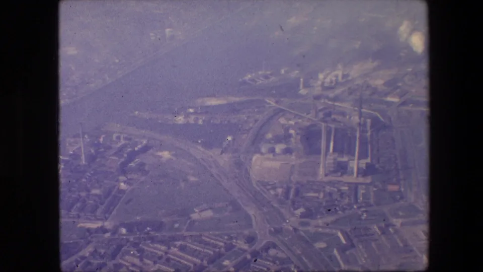 Aerial 8mm frame from the supplied 1967 New Jersey clip showing highway approaches crossing near a wide waterway, with industrial structures and smokestacks visible through haze.