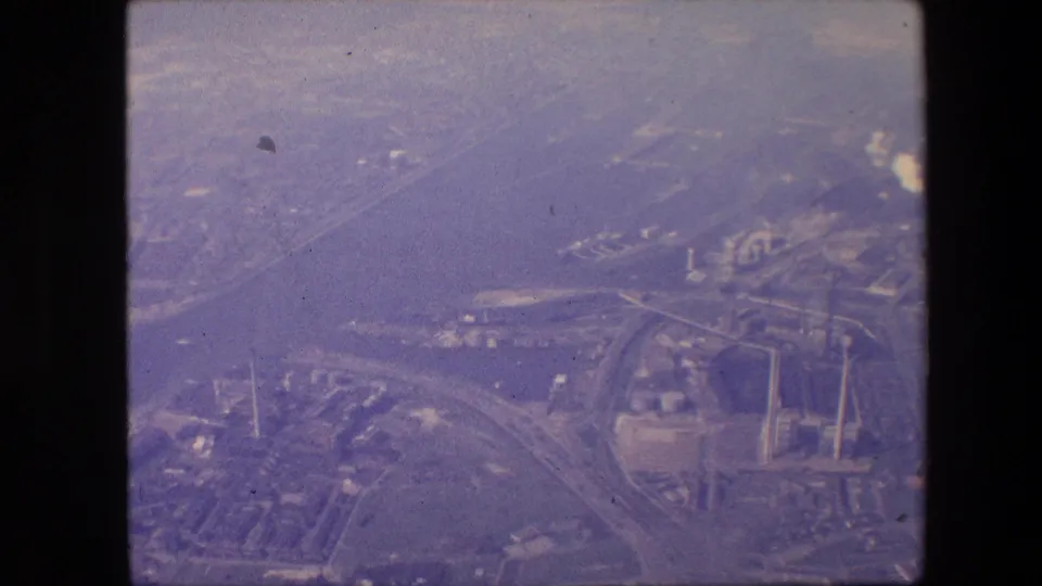 Soft purple-tinted 8mm aerial view, supported by the clip metadata as New Jersey in 1967, with a wide waterway, crossing roadways, industrial buildings, and nearby urban blocks.
