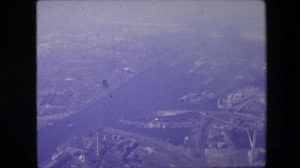 Final 8mm aerial frame from the supplied 1967 New Jersey footage, showing a hazy blue-purple view across a broad waterway toward industrial buildings, roads, and distant urban development.