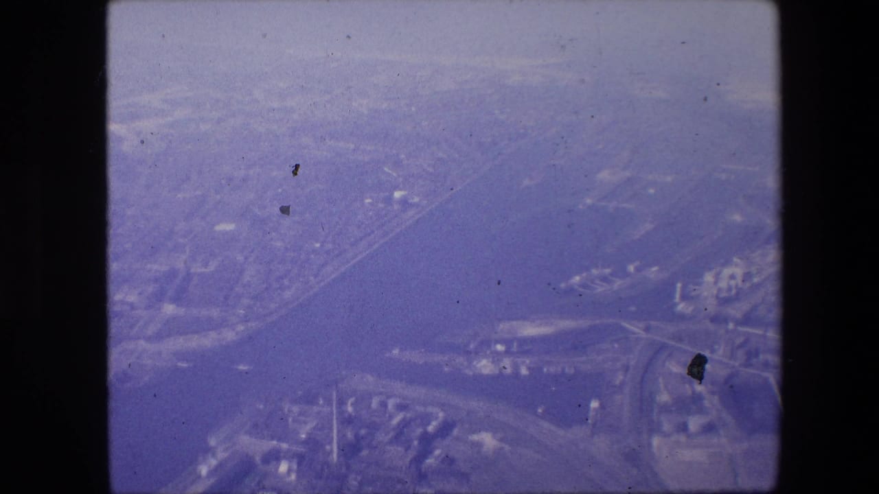 Aerial 8mm view associated with Amsterdam, showing a broad waterway, shoreline development, roads, and dense urban areas beneath a hazy purple film cast.