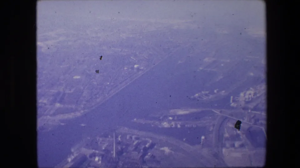 Aerial 8mm view associated with Amsterdam, showing a broad waterway, shoreline development, roads, and dense urban areas beneath a hazy purple film cast.