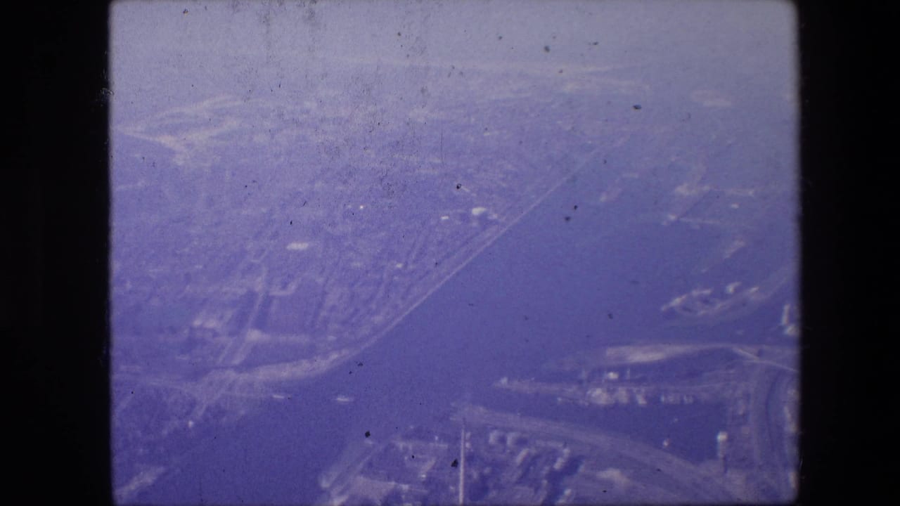 High-angle 8mm frame associated with Amsterdam, with a wide water channel, built-up shoreline, road network, and distant city fabric softened by haze and film grain.