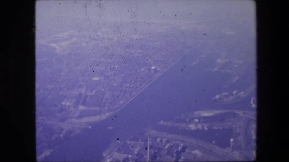 High-angle 8mm frame associated with Amsterdam, with a wide water channel, built-up shoreline, road network, and distant city fabric softened by haze and film grain.