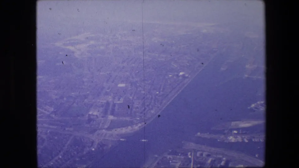 Oblique airplane-window view from 1967 metadata, showing an urban waterfront with roads, possible bridges, shoreline buildings, and soft purple 8mm film texture.
