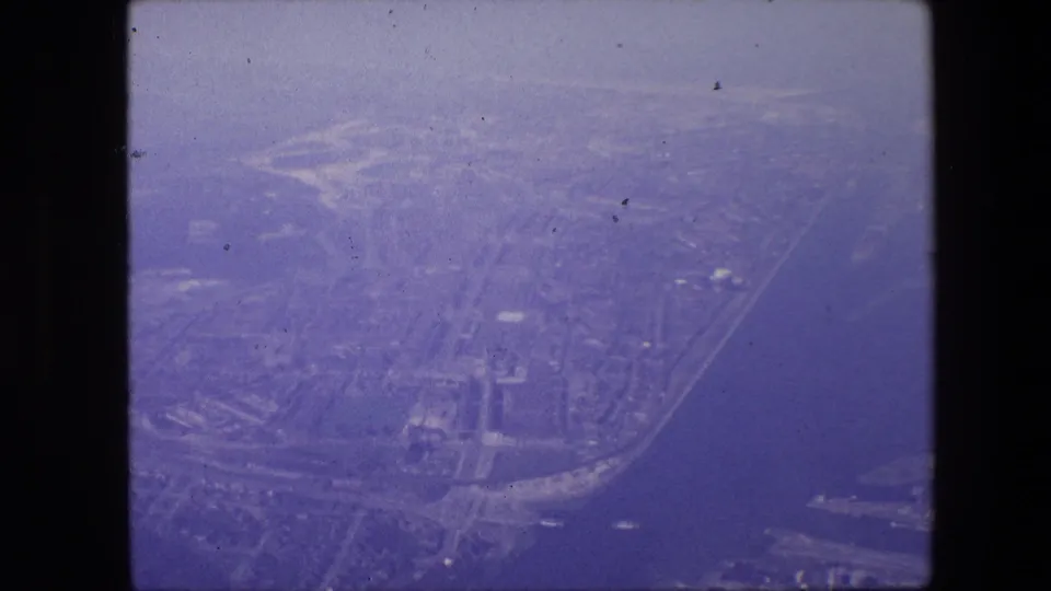 Vintage 8mm aerial frame associated with Amsterdam, showing a waterway cutting beside dense development, road corridors, shoreline infrastructure, and visible dust and grain from the film scan.