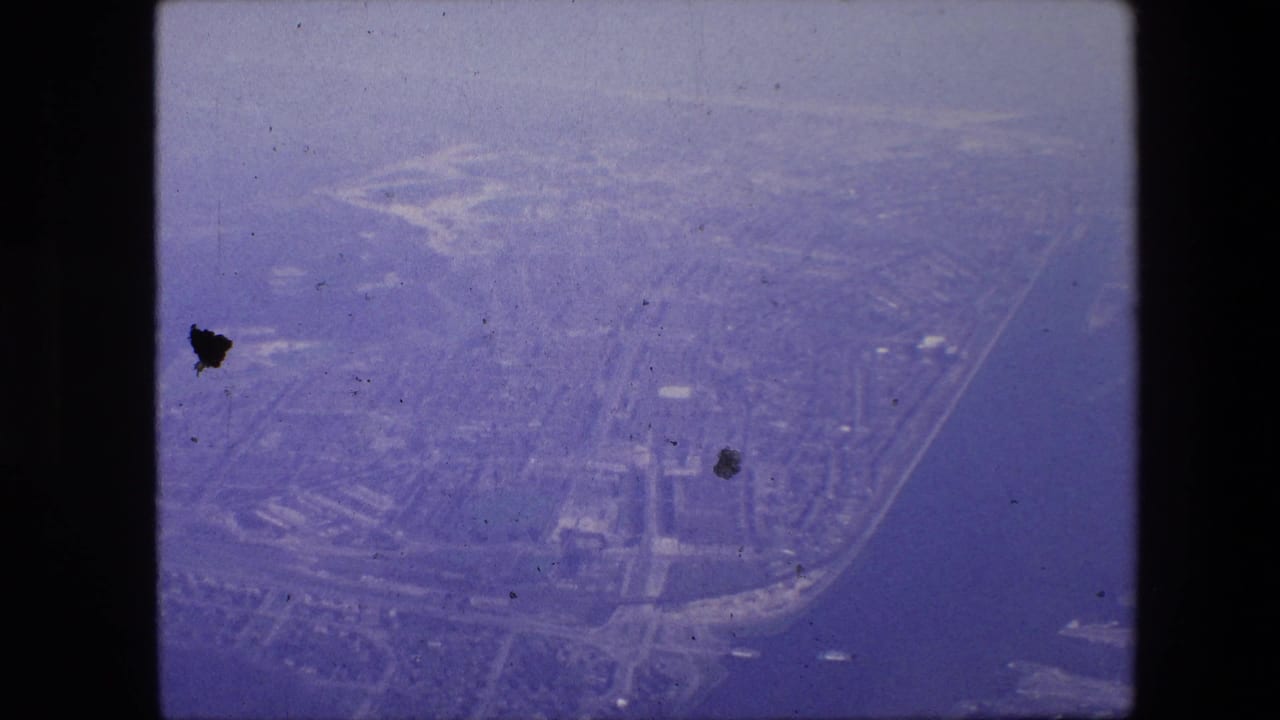 Hazy 1967 8mm aerial view over Amsterdam, showing canals or waterways, roads, rooftops, and shoreline forms under a purple-blue film cast.