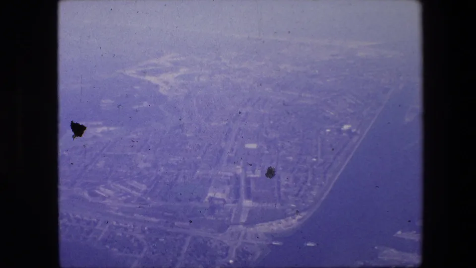 Hazy 1967 8mm aerial view over Amsterdam, showing canals or waterways, roads, rooftops, and shoreline forms under a purple-blue film cast.