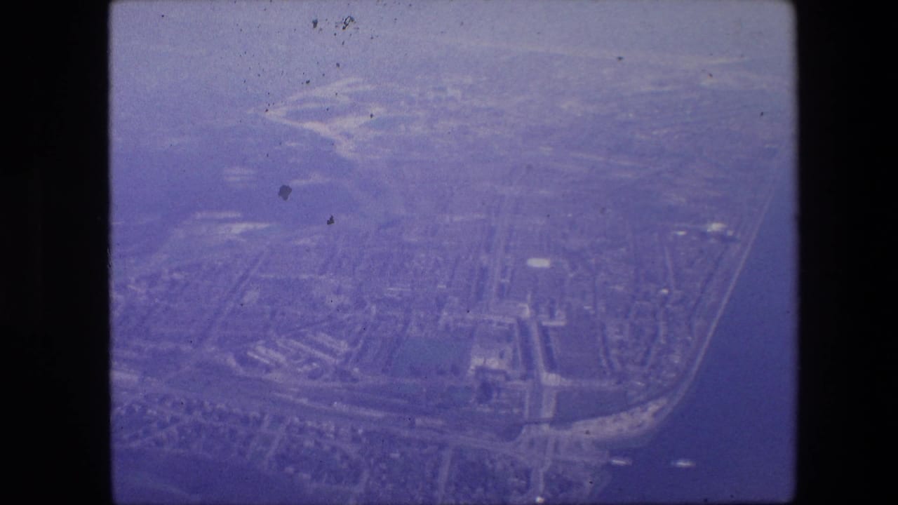 Aerial 8mm frame over Amsterdam in 1967, with a dense urban pattern, canals, road lines, and soft haze visible from above.