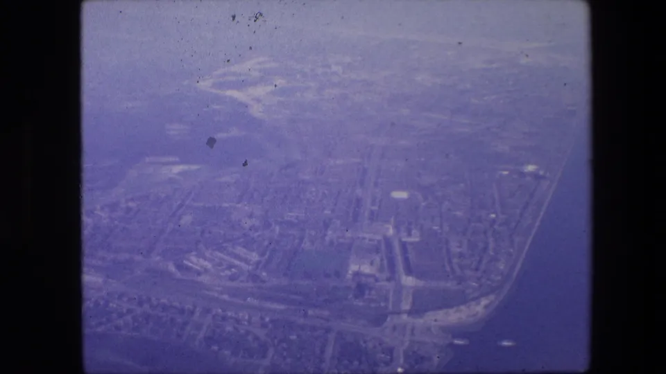 Aerial 8mm frame over Amsterdam in 1967, with a dense urban pattern, canals, road lines, and soft haze visible from above.