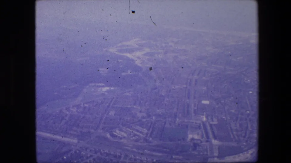 Soft 1967 aerial home-movie view of Amsterdam, with canal-like water channels, clustered buildings, and broad road corridors visible through faded color.