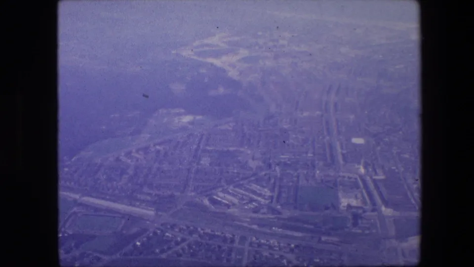 Purple-tinted 8mm aerial view over Amsterdam in 1967, showing a canal network, bridges, roadways, rooftops, and waterfront edges from above.