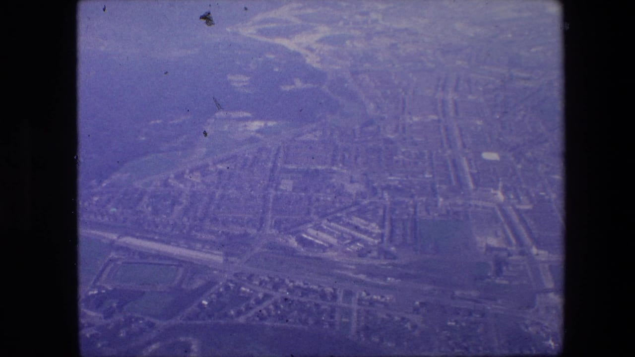1960s 8mm-style aerial view from an airplane window showing a hazy urban grid below, with no identifiable landmark visible in the frame.